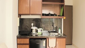 A compact kitchen with wooden cabinets, a dishwasher, a coffee maker, and a green cutting board on the countertop. - Nice and Comfort 1BR with Extra Room at Podomoro Golf View Apartment (Gunung Putri)