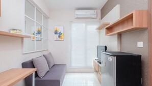 Un espace de vie compact comprenant un canapé, une petite table, un réfrigérateur et une fenêtre avec des stores.