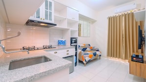 A compact kitchen with a built-in gas stove, a sink, and a compact refrigerator. There is a bed with a colorful blanket and a small bedside table with a bottle of water and a travel pillow. - Comfort Podomoro Golf View 2BR Apartment (Gunung Putri)