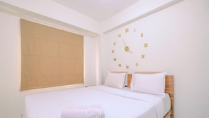A neatly made bed with white linens and a folded towel, a wooden headboard, and a wall clock with Roman numerals. - Comfort Podomoro Golf View 2BR Apartment (Gunung Putri)