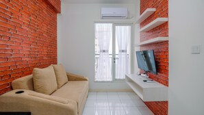Un petit salon moderne avec un mur en briques, un canapé beige, un téléviseur à écran plat posé sur une étagère blanche et une porte-fenêtre coulissante avec des rideaux légers.