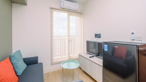 A compact living space with a sofa, a small table, a refrigerator, a television, and a window with an air conditioning unit. - Minimalist and Warm 2BR at 29th Floor Podomoro Golf View Apartment (Gunung Putri)