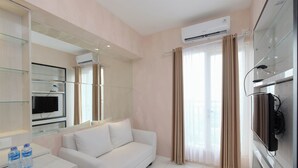 A hotel room with a white sofa, a TV mounted on the wall, a glass cabinet, and an air conditioning unit. - Comfort and Stylish 2BR at Podomoro Golf View Apartment (Gunung Putri)