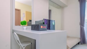 A modern hotel room with a white desk, a small potted plant, a minibar, and a bed with a beige cover. - Best Deal and Tidy 2BR Podomoro Golf View Apartment (Gunung Putri)