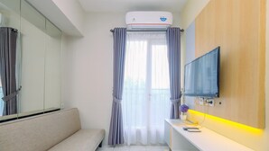 A modern hotel room with a sofa, a TV mounted on the wall, a white desk with a lamp, and a large window with curtains. - Best Deal and Tidy 2BR Podomoro Golf View Apartment (Gunung Putri)