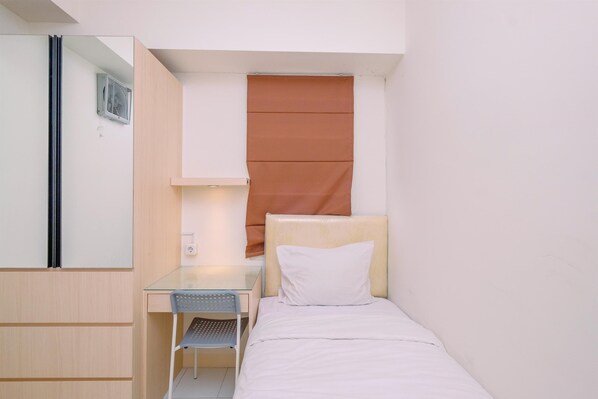 Una habitación pequeña y funcional con una cama, un escritorio y una silla. Se encuentra un armario en un lateral y una ventana con persianas.