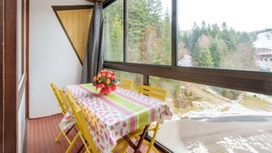 Ein Balkon mit Tisch und Stühlen, ein Fenster mit Blick auf Bäume und ein Gebäude, sowie eine Holzbalkendecke.