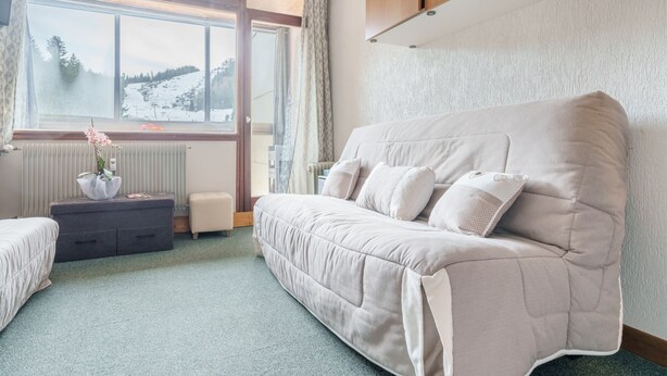 Ein gemütliches Zimmer mit großem Fenster, einer Couch, einem kleinen Tisch mit einer Vase und einem Heizkörper.