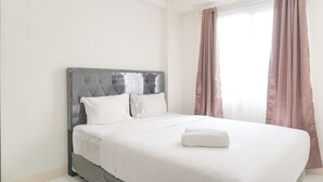 A neatly made bed with white linens and a folded towel, a grey upholstered headboard, and a window with brown curtains. - Comfy Stay 2BR at Podomoro Golf View Apartment (Gunung Putri)