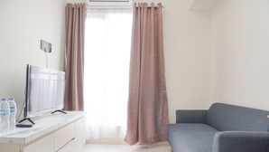 A modern living room with a flat-screen TV on a white console, a grey sofa, and a window with sheer curtains. - Comfy Stay 2BR at Podomoro Golf View Apartment (Gunung Putri)