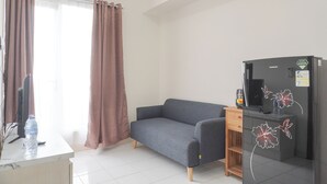 A modern room with a grey sofa, a wooden desk, a black refrigerator, and a window with curtains. - Comfy Stay 2BR at Podomoro Golf View Apartment (Gunung Putri)