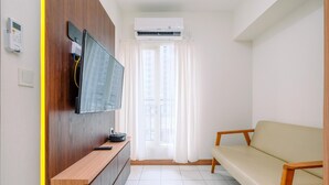 A modern living room with a flat-screen TV mounted on a wooden wall, a sofa, a wooden cabinet, and a window with white curtains. - Tidy and Comfortable 2BR Podomoro Golf View Apartment (Gunung Putri)