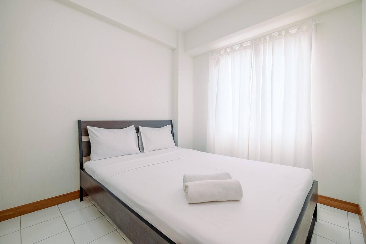 A neatly made bed with white linens and a folded towel, a wooden headboard, and a window with sheer curtains.