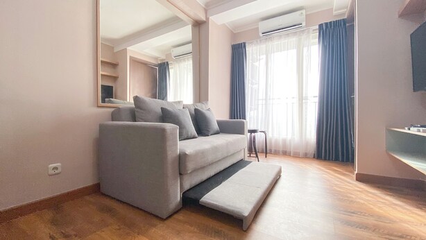 A modern living room with a grey sofa, a small side table, and a bench. There is a large mirror on the wall, a window with curtains, and an air conditioning unit above the window. - Best Deal and Homey 3BR Podomoro Golf View Apartment (Gunung Putri)