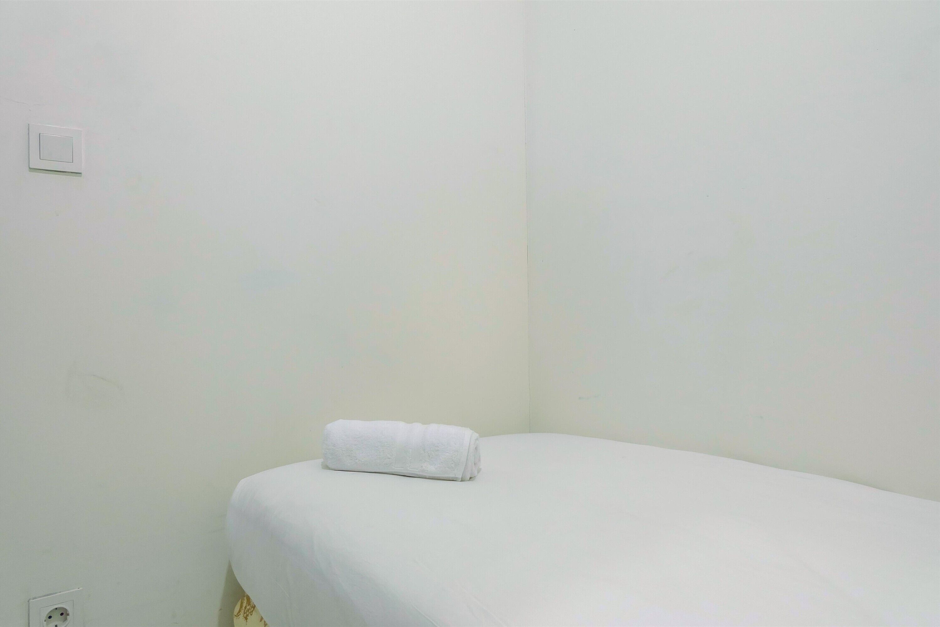 Ein Einzelbett mit einer ordentlich gefalteten weißen Decke in einem Zimmer mit schlichten weißen Wänden und einer sichtbaren Steckdose.