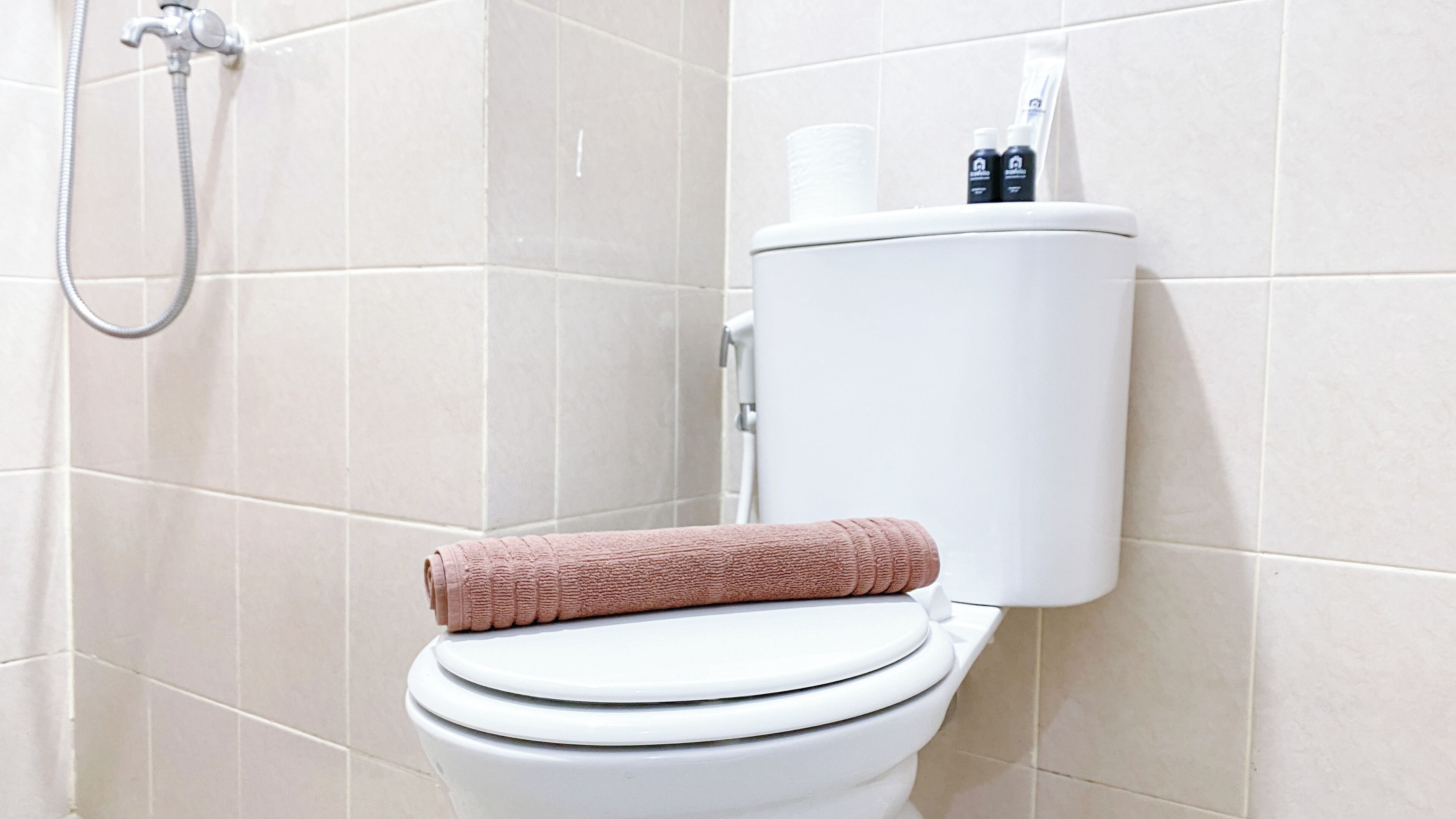 Des toilettes avec une serviette rose posée sur le siège, un porte-rouleau de papier toilette et une pomme de douche.