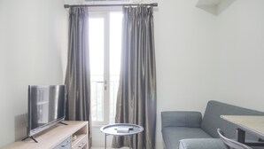 A modern living room with a flat-screen TV on a wooden stand, a grey sofa, a small round table, and a window with grey curtains. - Best Choice and Homey 2BR Podomoro Golf View Apartment (Gunung Putri)