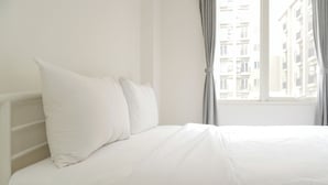 Ein weißes Bett mit Kopfteil, ein Fenster mit Vorhängen und ein Blick auf Gebäude im Freien.