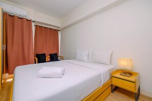 A neatly made bed with a white comforter and a rolled white towel, a bedside table with a lamp, and a window with closed curtains.