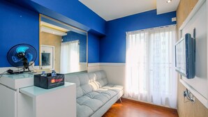 Une pièce avec un mur bleu, un canapé gris, un bureau blanc, un ventilateur noir, un miroir, un téléviseur et une fenêtre avec des rideaux blancs.