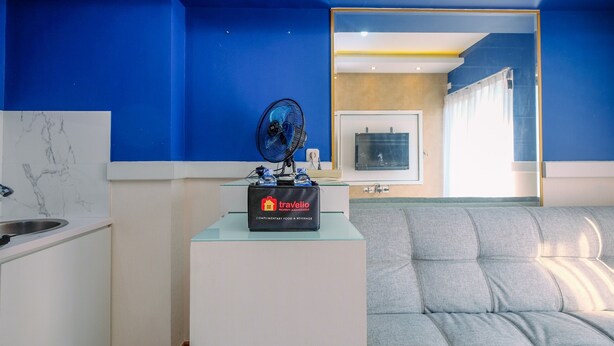 A modern hotel room with a blue accent wall, a grey sofa, a glass coffee table with a minibar, and a large mirror reflecting the room. - Comfy and Tidy 1BR at Podomoro Golf View Apartment (Gunung Putri)