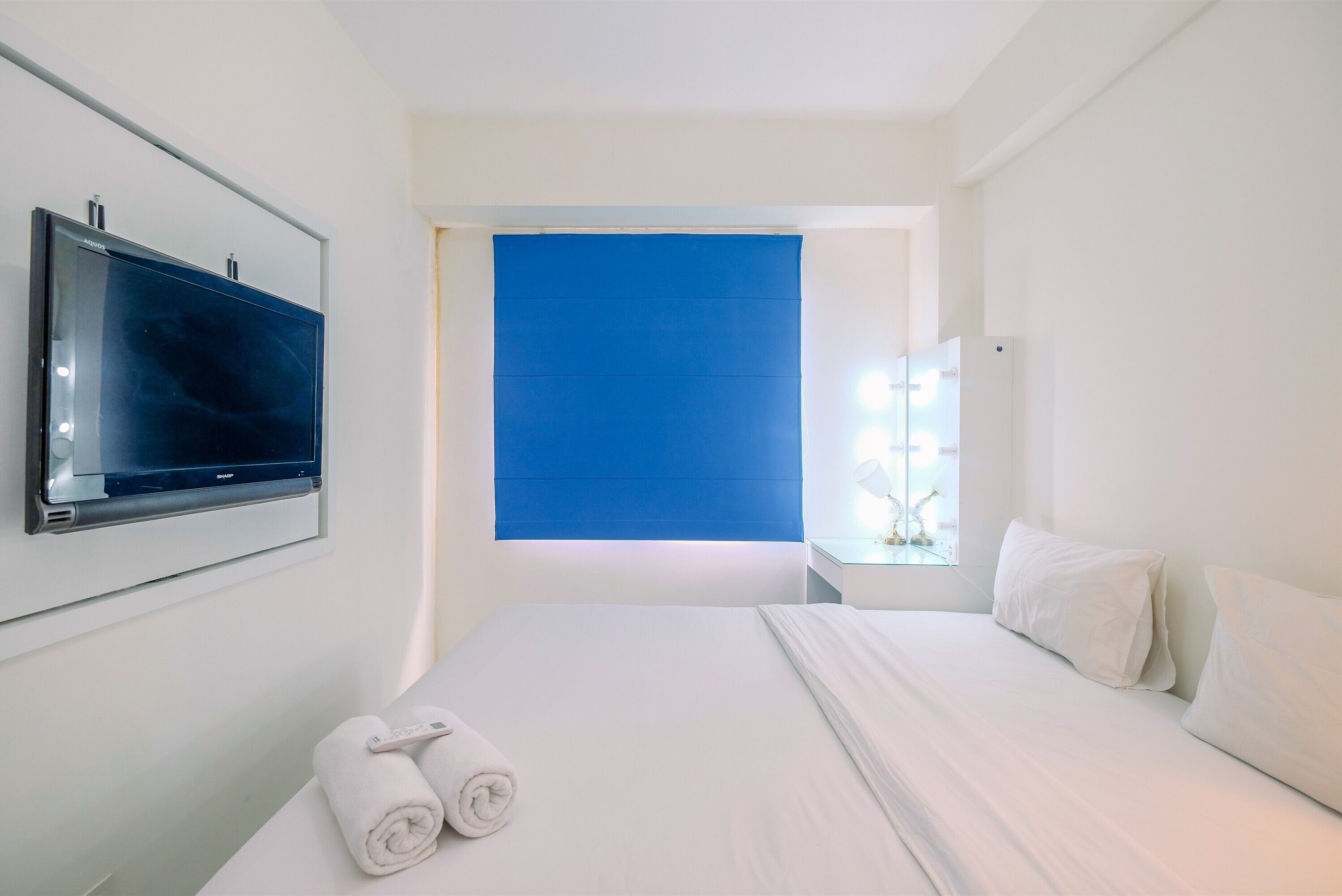 Ein Hotelzimmer mit einem Bett, einem an der Wand befestigten Fernseher, einem Fenster mit blauer Jalousie und einem kleinen Tisch mit Lampe.