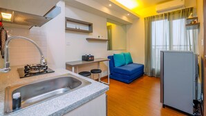 A compact kitchen with a sink, stove, and bar stools, a blue sofa, and a refrigerator in a modern apartment. - Minimalist and Cozy Stay 2BR at Podomoro Golf View Apartment (Gunung Putri)