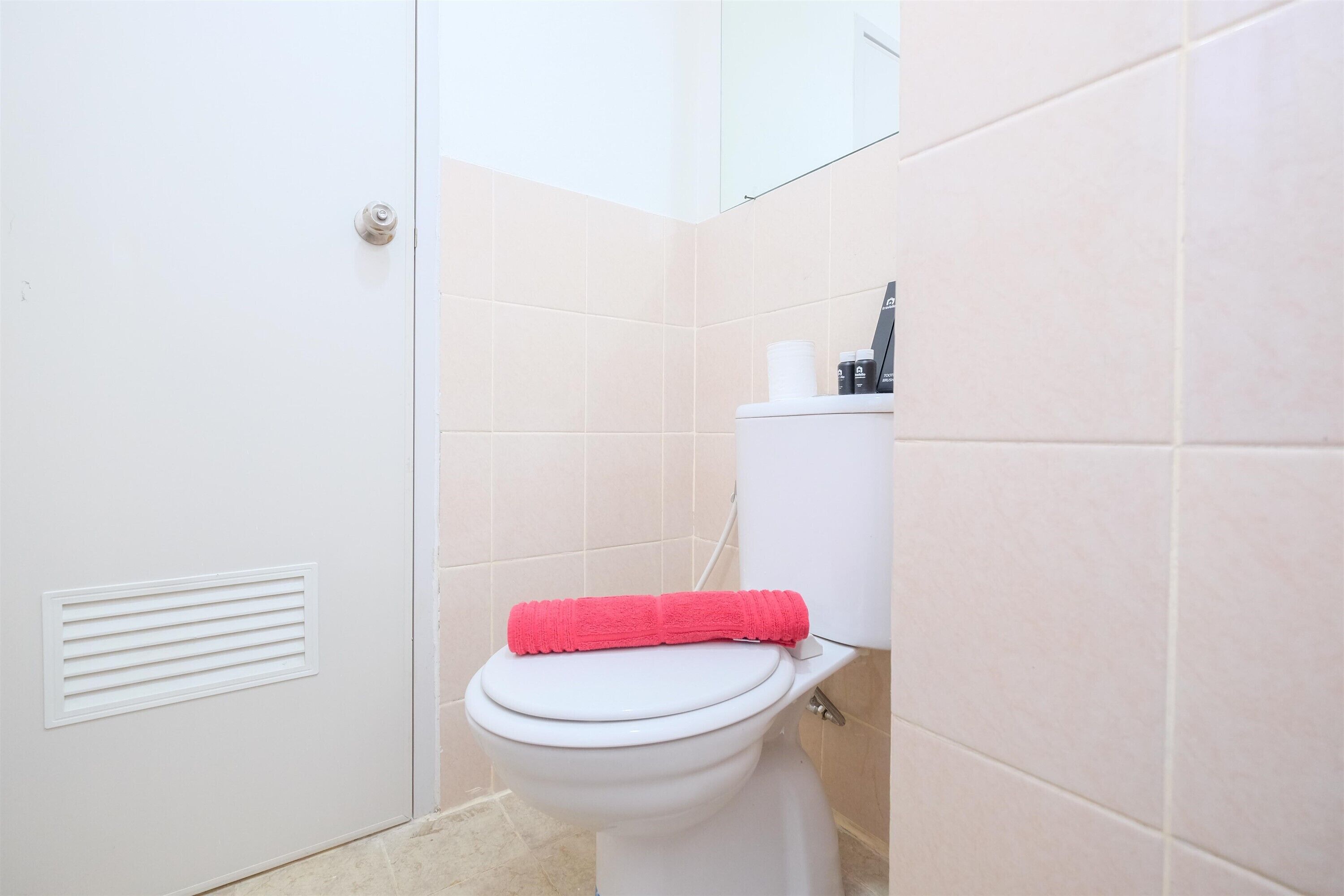 Une salle de bain avec des toilettes blanches, des carreaux beiges et une serviette rouge posée sur le couvercle.