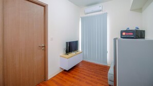 Une petite pièce bien éclairée, avec une porte en bois, un meuble blanc contenant un téléviseur à écran plat, un réfrigérateur et une fenêtre munie de stores.