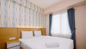 A neatly made bed with white linens and a folded towel, a wooden nightstand with a power outlet, and a window with blue curtains. - Comfort and Restful 2BR Podomoro Golf View Apartment (Gunung Putri)