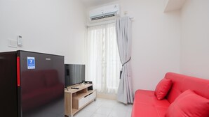 A compact living space with a black refrigerator, a red sofa, a wooden TV stand, and a window with white curtains. - Comfort 2BR at 6th Floor Podomoro Golf View Apartment (Gunung Putri)