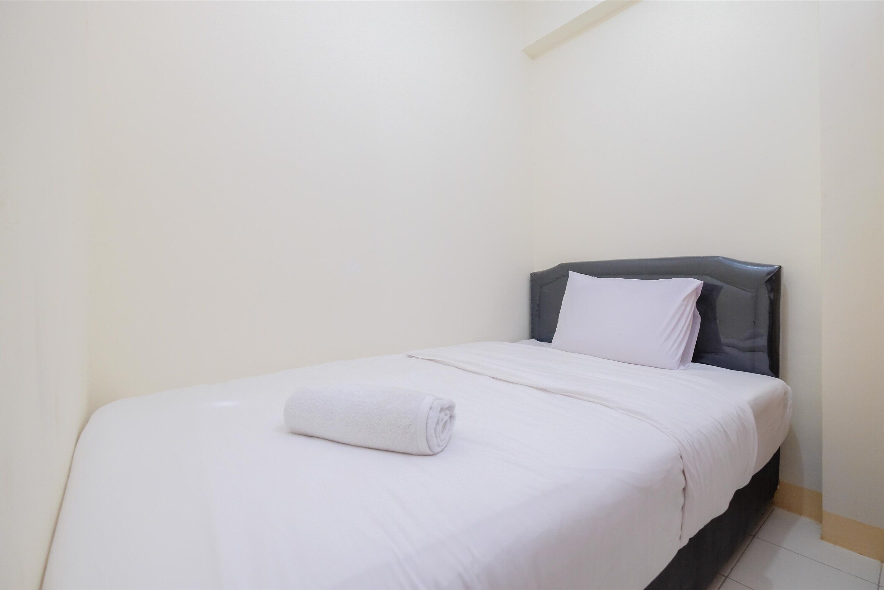 A neatly made bed with white linens and a rolled towel, a gray headboard, and a plain white wall.