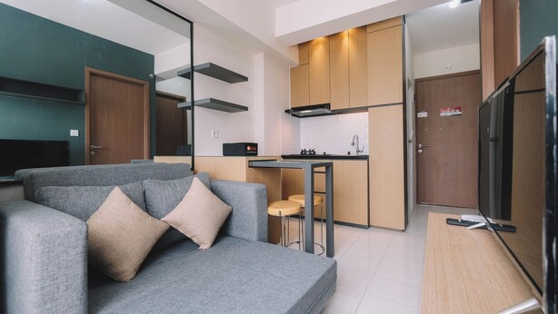 A modern living room with a grey sofa, a TV, and a kitchen area with wooden cabinets and a small bar counter. - Simple and Cozy Living 2BR at Podomoro Golf View Apartment (Gunung Putri)