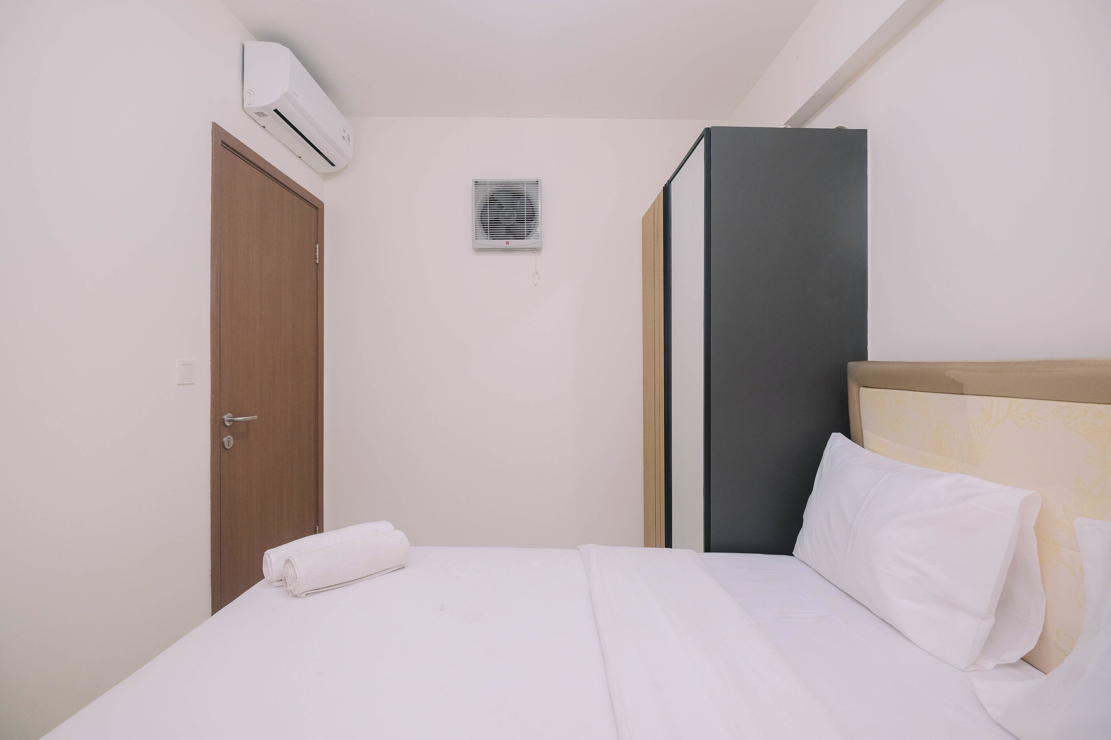 A neatly made bed with white linens, a wooden door, and a wall-mounted air conditioner.