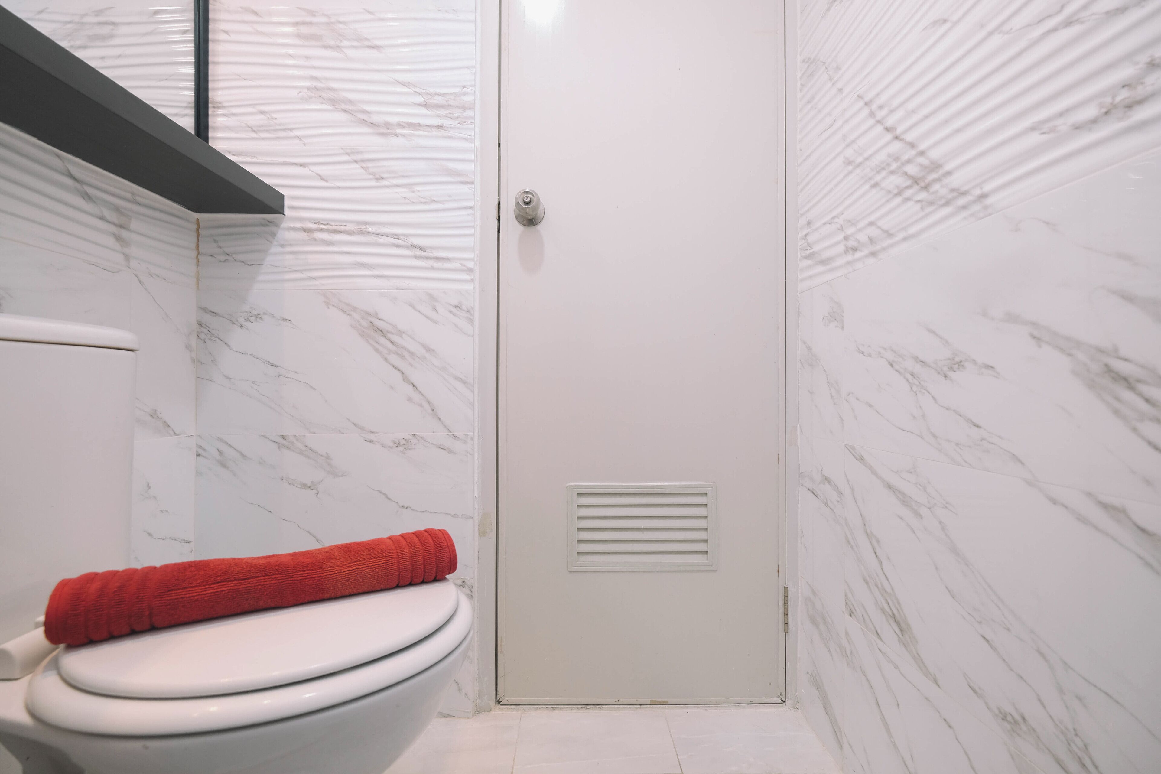 A bathroom with a white toilet, a red towel on the seat, and a door with a vent.