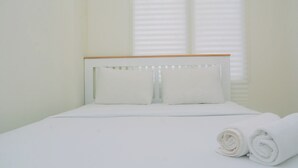 Een netjes opgemaakt bed met witte lakens en kussens, een houten hoofdbord en een raam met jaloezieën.