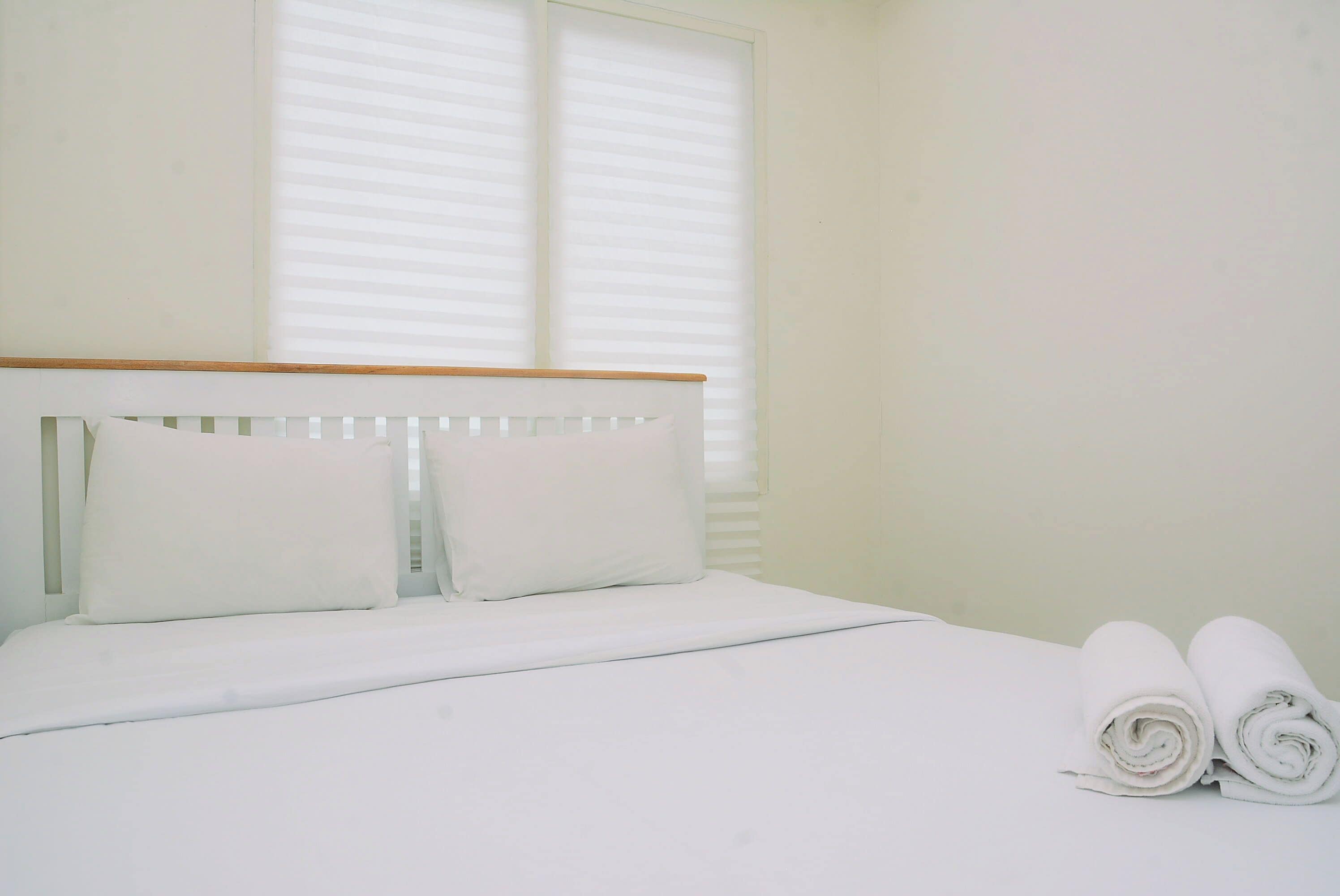 A neatly made bed with white linens and pillows, a wooden headboard, and two rolled towels on the side.