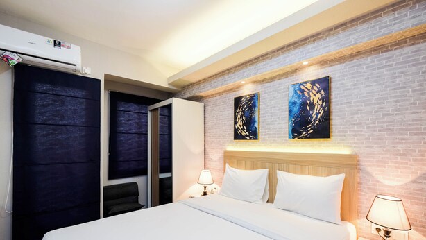 A hotel room with a bed, two bedside tables with lamps, a wardrobe, and a brick wall with two artworks. - Brand New and Fresh Studio Podomoro Golf View Apartment (Gunung Putri)