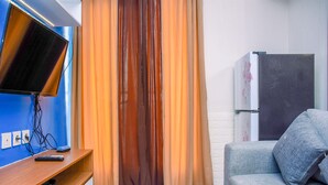 A room with a TV on a wooden stand, a sofa, a refrigerator, and curtains. - Nice and Warm 2BR at Podomoro Golf View Apartment (Gunung Putri)