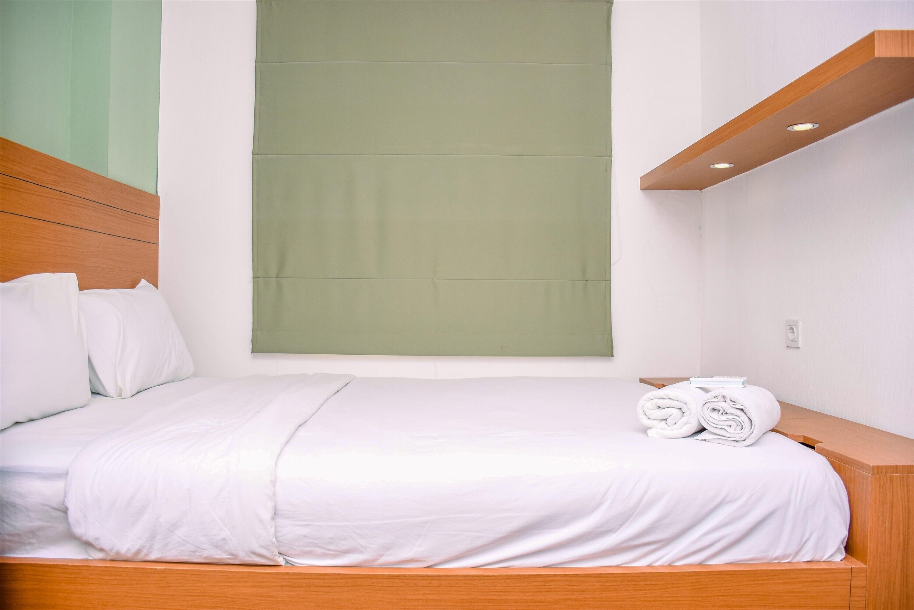 A neatly made bed with white linens and a wooden headboard, a green window blind, and a wooden bedside table with rolled towels.