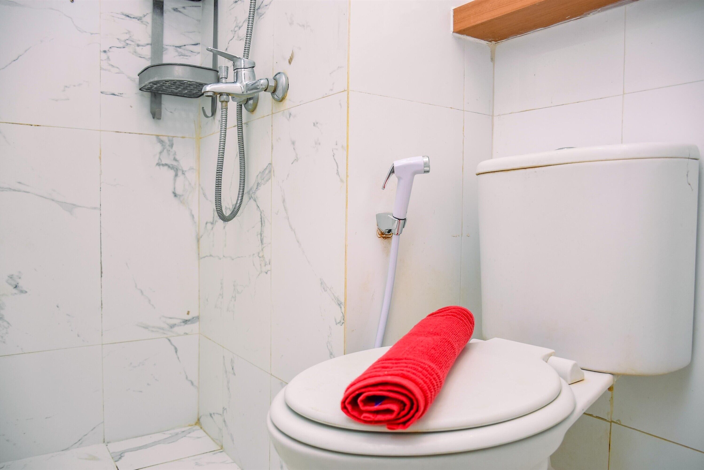 A bathroom with a white toilet, a red towel on the seat, and a shower head.