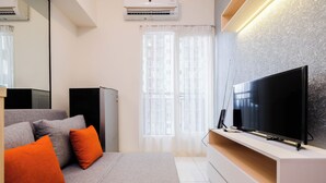 A modern living room with a large flat-screen TV, a sofa with orange cushions, a small cabinet, and a window with sheer curtains.