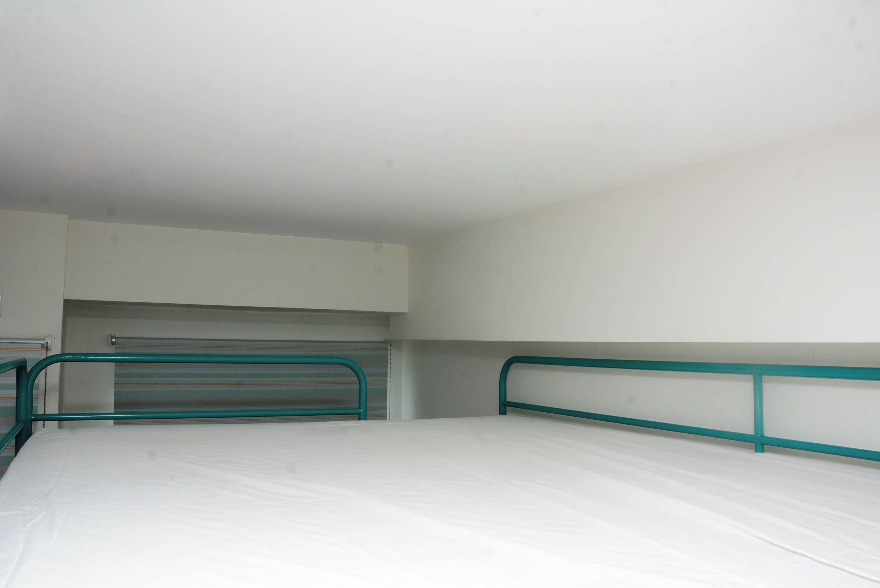 A single-bed room with a metal frame, white walls, and a small window.