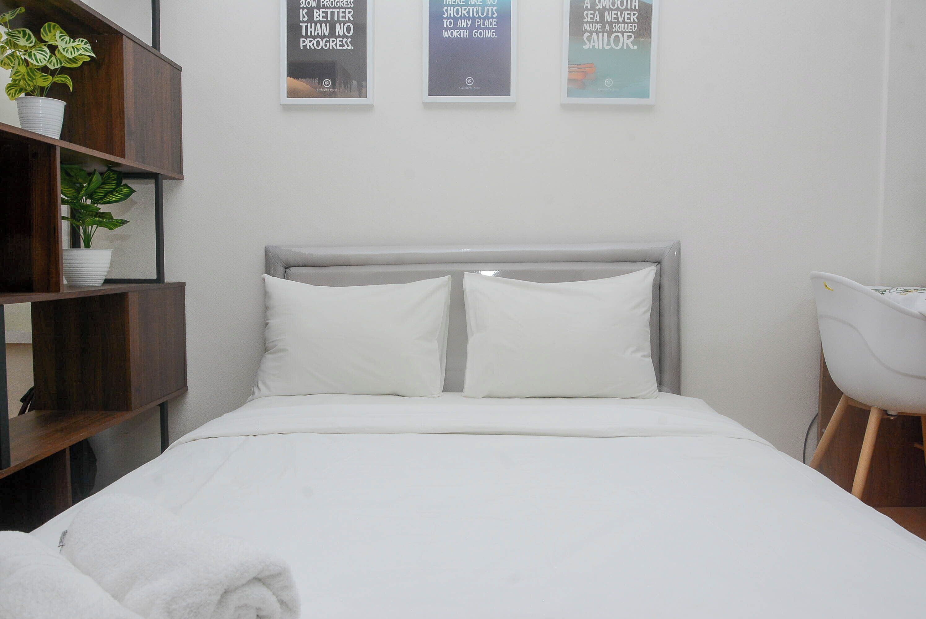 Una cama bien hecha con ropa de cama blanca, un cabecero gris y dos almohadas blancas. Hay tres carteles motivacionales enmarcados en la pared sobre la cama.