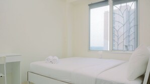 A clean, white room with a single bed, a small desk, and a window with a view of buildings.