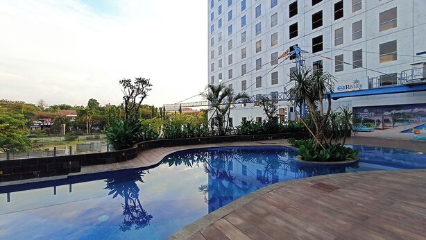 A swimming pool with a wooden deck, surrounded by greenery and a modern building with multiple windows. - Brand New and Homey Studio at Tamansari Skylounge Makassar Apartment (Makassar)