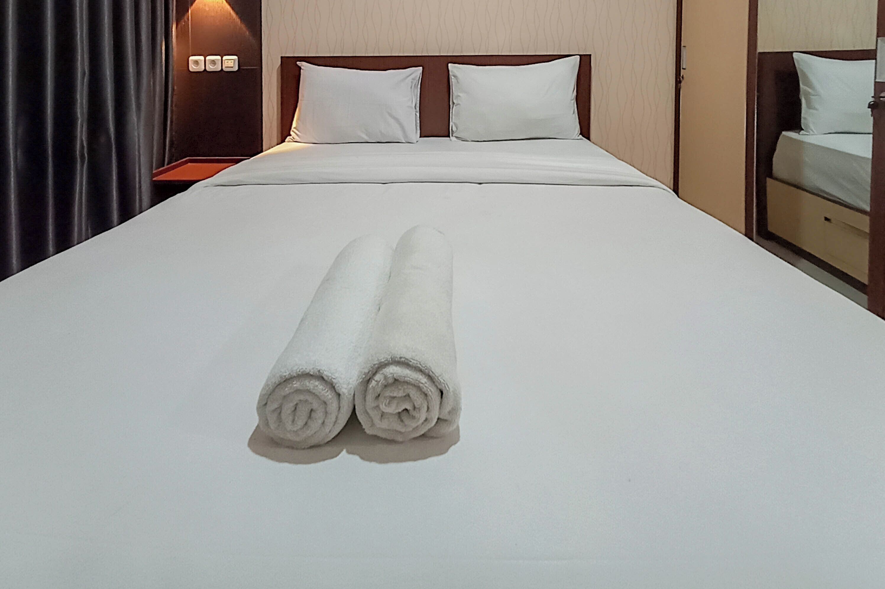 A neatly made bed with two rolled white towels in front, a wooden headboard, and a nightstand with a lamp.