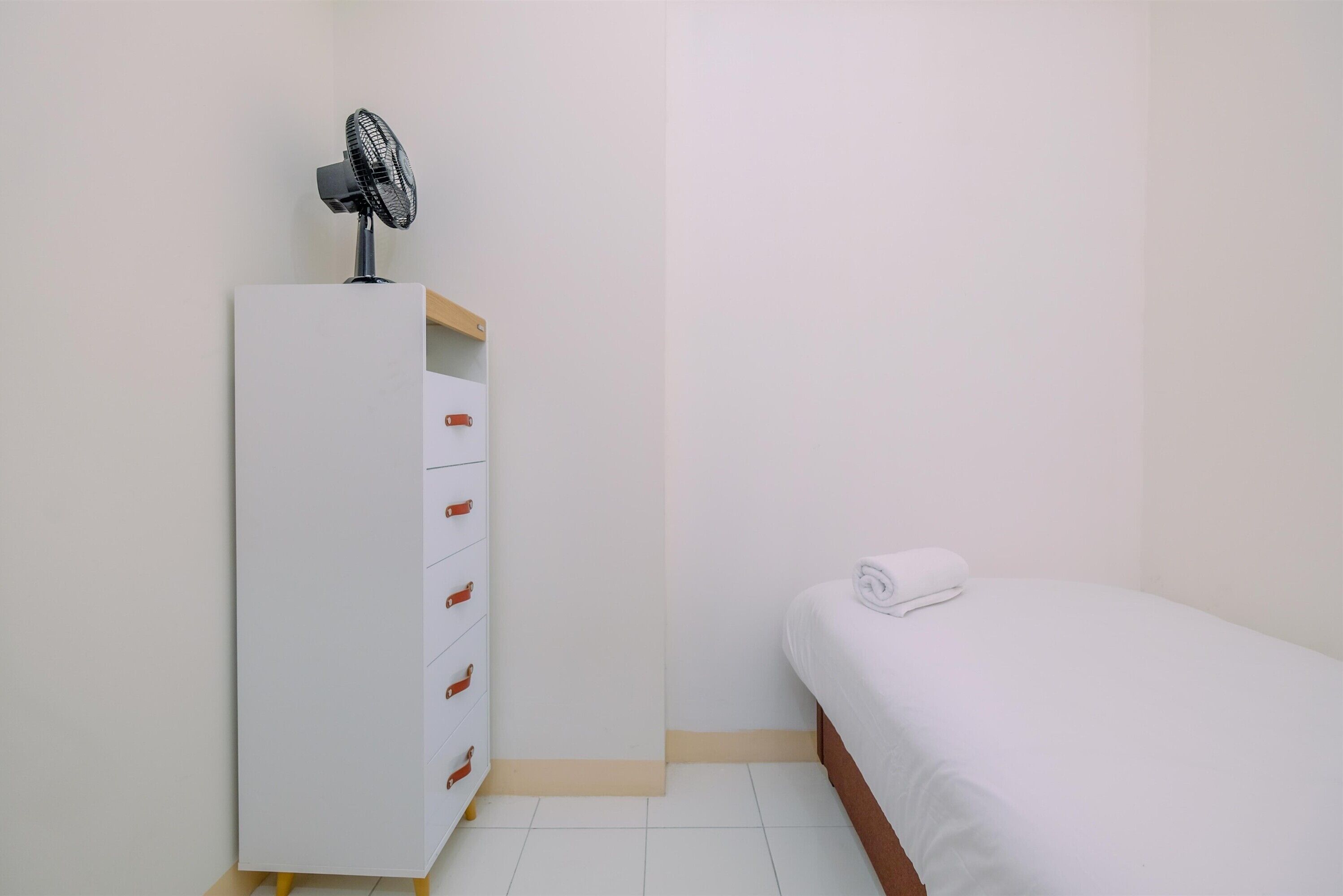 A bedroom with a white bed, a white wardrobe with drawers, and a standing fan.