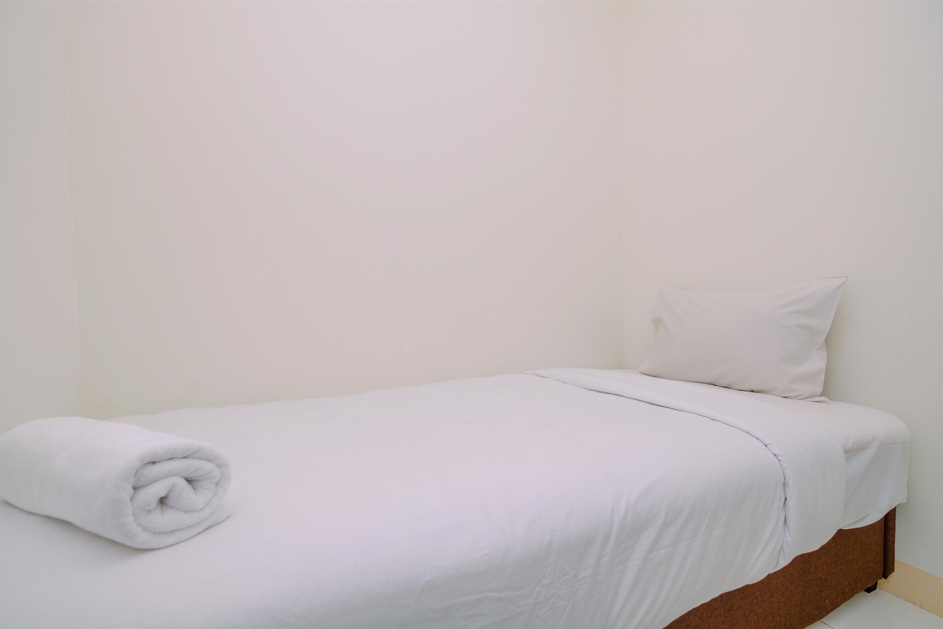 Ein Einzelbett mit weißer Matratze, einem aufgerollten weißen Handtuch, einem weißen Kissen und einem braunen Kopfteil in einem Raum mit schlichten weißen Wänden.