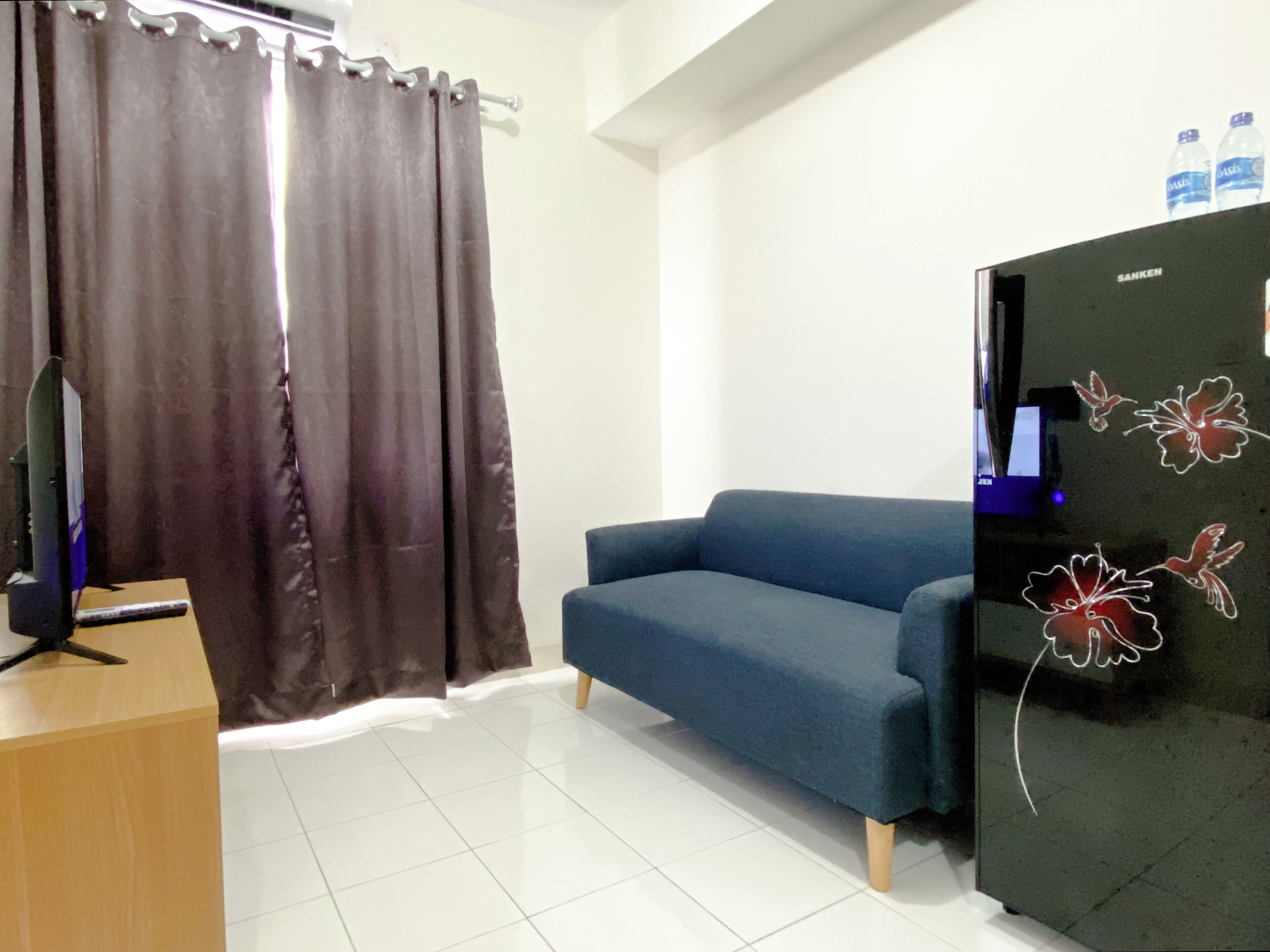 Una sala de estar con un sofá azul, una mesita de madera, un refrigerador negro con botellas de agua y cortinas marrones.